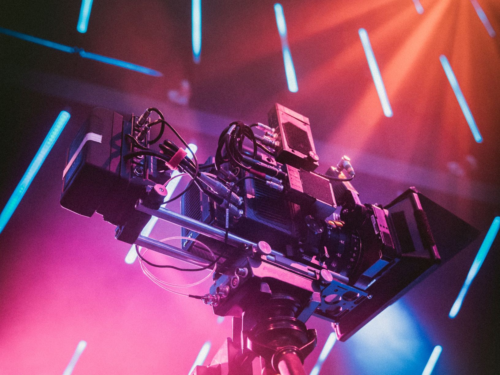 A camera is pictured. The background lights are pink, orange and blue. The atmosphere is dust-laden and rays of lights are visible. There are parallel white-blue stripes in the background.